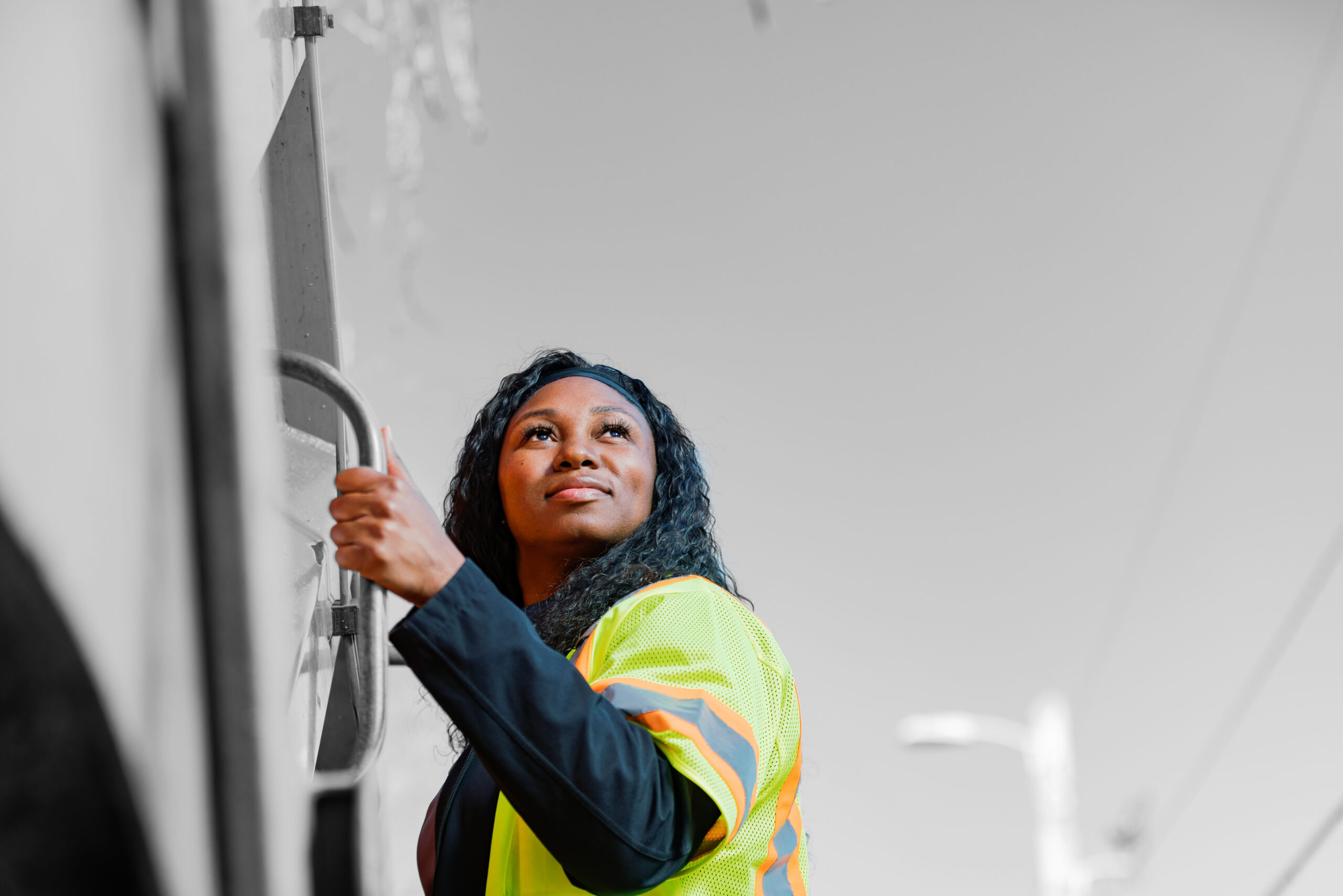 A woman wearing a yellow safety vest stands outdoors, looking upward and holding onto a metal handle, with a blurred background in grayscale.