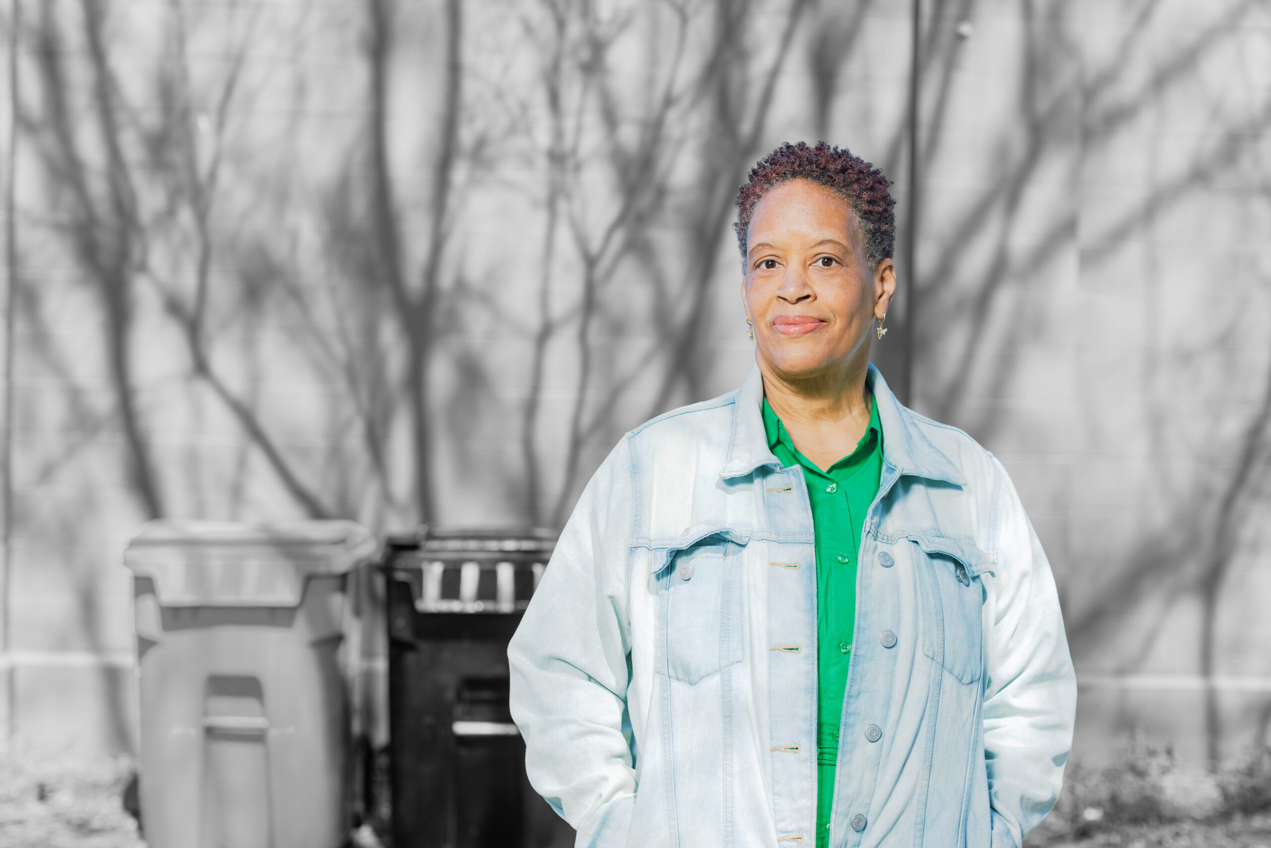 A woman with short curly hair, wearing a light denim jacket over a green shirt, stands outdoors in front of trash bins and a wall with tree shadows on it.