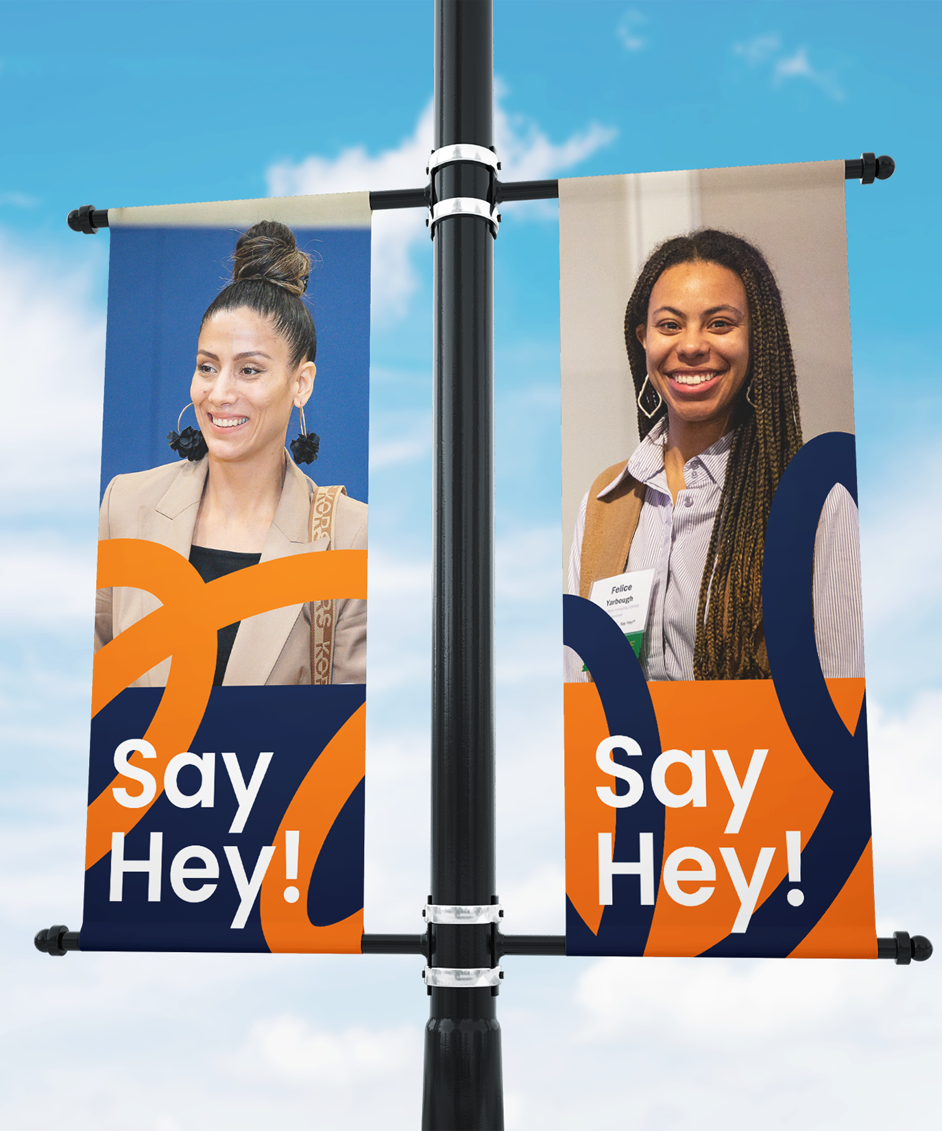 Two street banners on a pole show smiling women in business attire with abstract orange and blue designs. Each banner reads Say Hey! against a bright sky background.