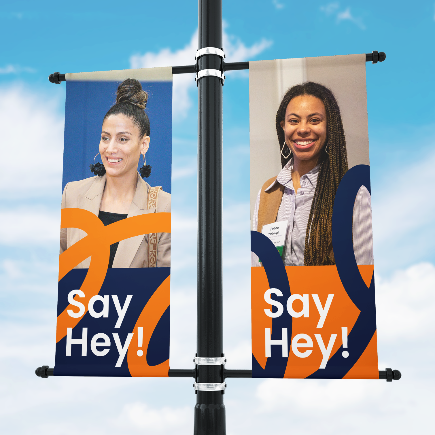 Two outdoor banners on a pole feature portraits of two women smiling, with abstract orange and blue designs and the words Say Hey! on both banners against a blue sky with clouds.
