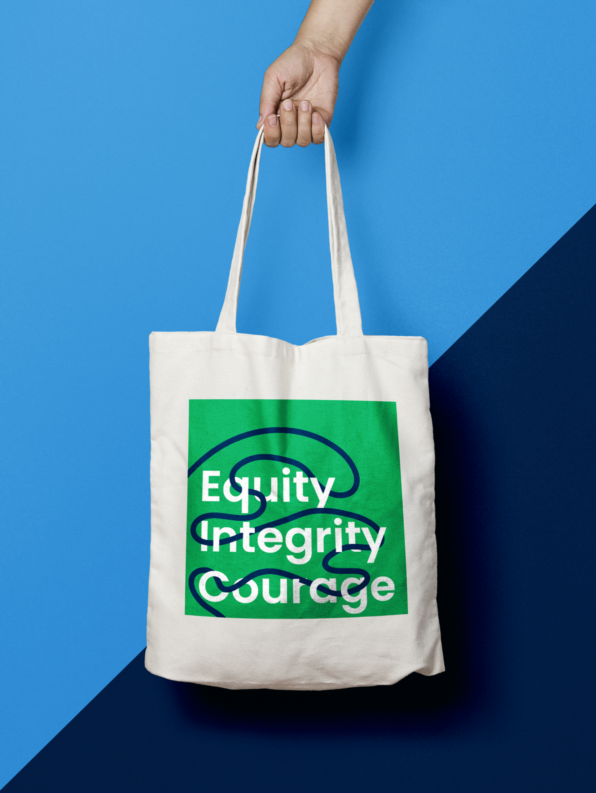 A hand holds a white tote bag with a green square design featuring the words Equity Integrity Courage and abstract blue lines on a blue and navy background.