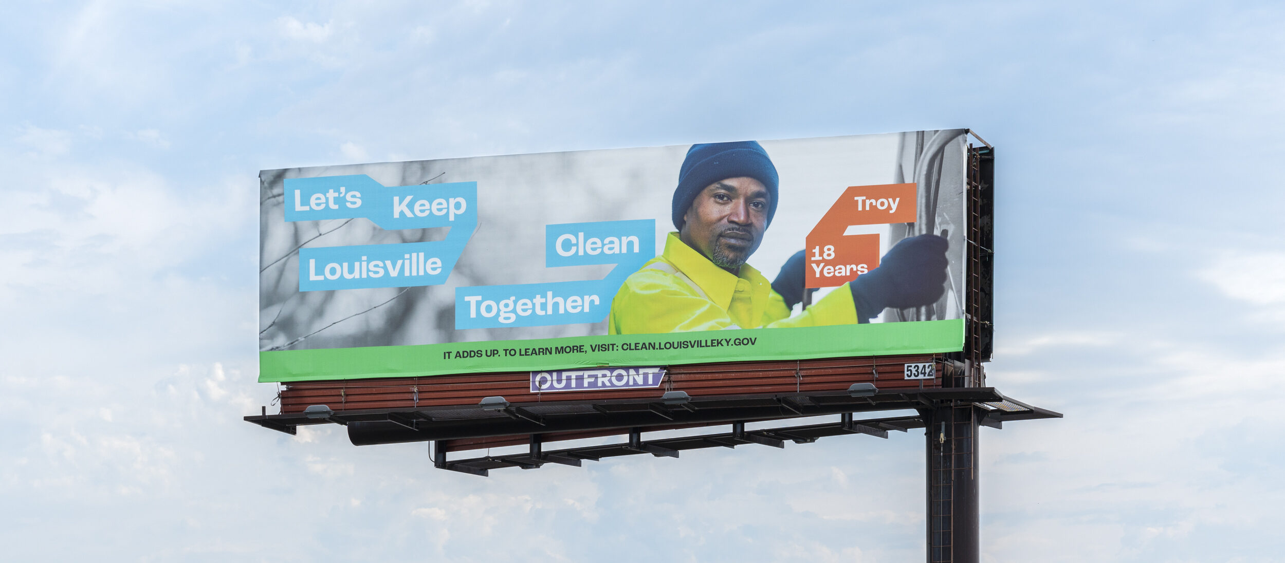 A billboard shows a man in a yellow jacket and blue beanie with text: Lets Keep Louisville Clean Together and Troy, 18 Years. The sky is blue with light clouds in the background.