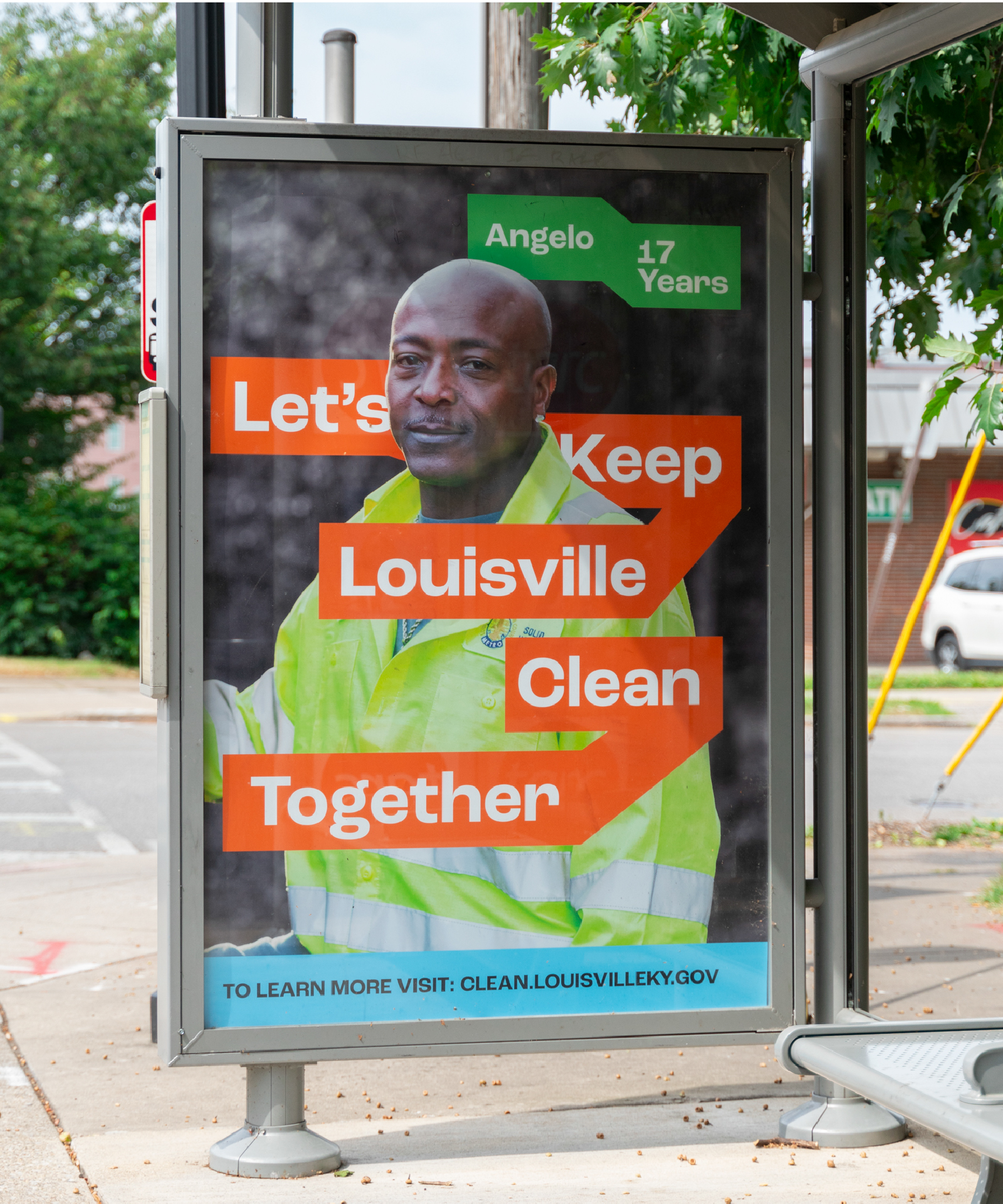 A bus stop ad shows a man in a yellow safety jacket with text: Angelo, 17 Years. Let’s Keep Louisville Clean Together. The background includes trees and a street. The ad promotes clean.louisvilleky.gov.