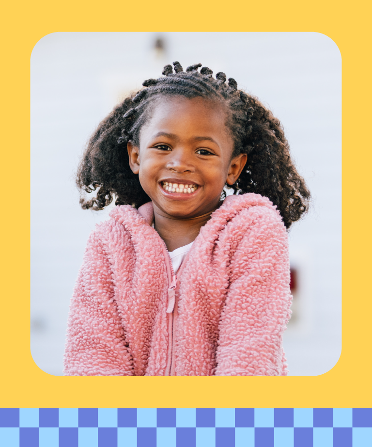 A young girl with curly hair and braids smiles brightly, wearing a pink fuzzy jacket. The photo has a yellow border with rounded corners and a blue checkered pattern along the bottom.