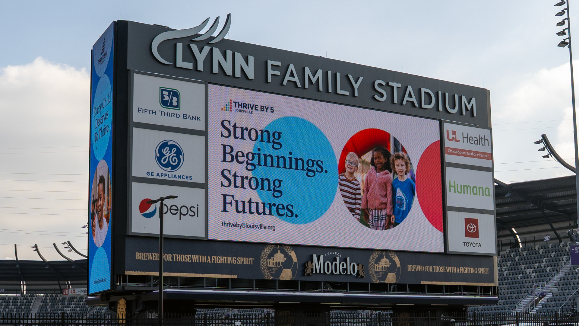 Large digital scoreboard at Lynn Family Stadium displays an ad with the text Strong Beginnings. Strong Futures. and three young children. Sponsor logos for local businesses are visible around the screen.