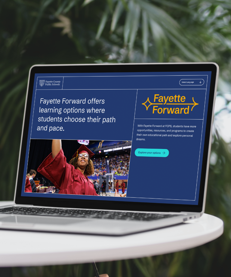 A laptop on a table displays a Fayette County Public Schools webpage about Fayette Forward, featuring a student in a graduation gown raising their arm in celebration at a graduation ceremony.