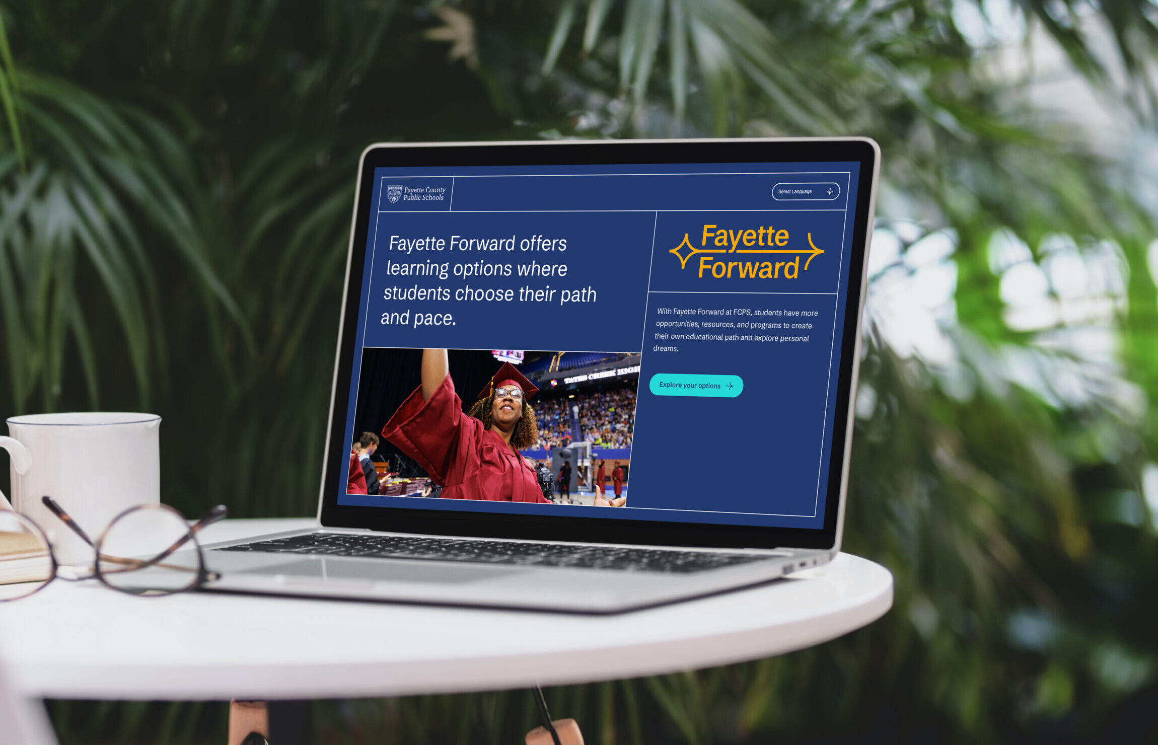 A laptop on a table displays the Fayette Forward website, featuring a photo of a smiling graduate in a cap and gown, and text about flexible learning options. The background is blurred greenery.