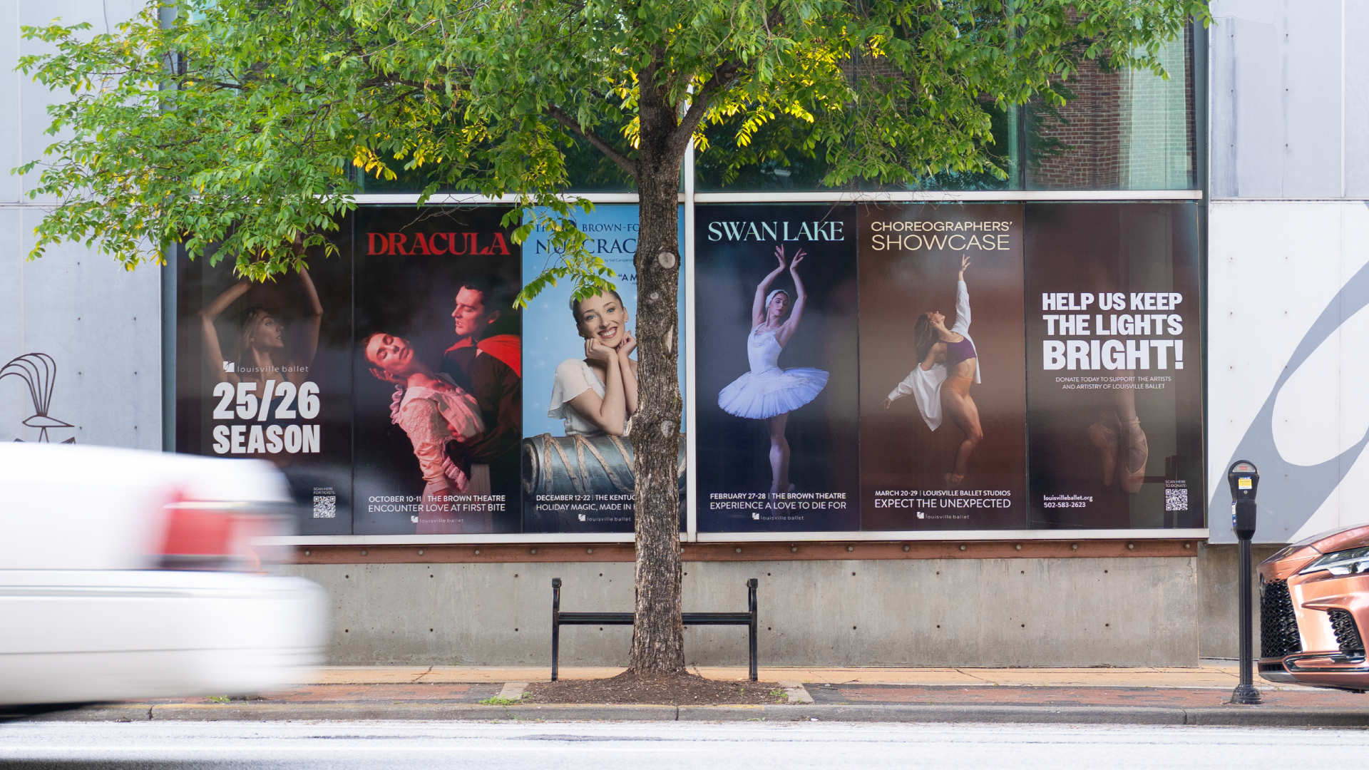 Four ballet posters are displayed in a row on a building wall behind a tree. A white car is blurred as it drives past, and a parked orange car is partially visible. The posters promote shows like Dracula, The Nutcracker, and Swan Lake.