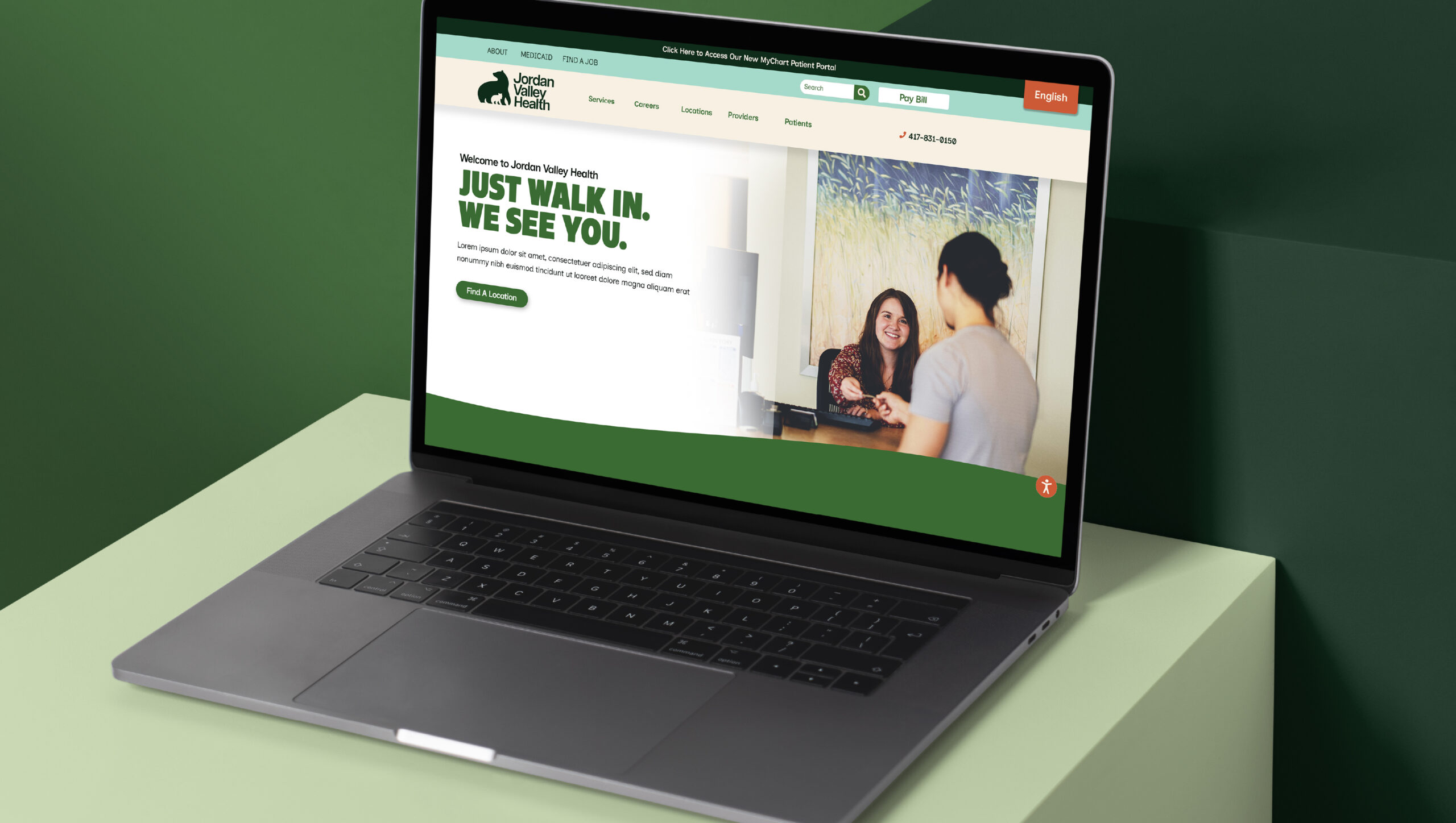 A laptop on a green surface displays a healthcare website showing one person talking to a receptionist at a clinic counter.
