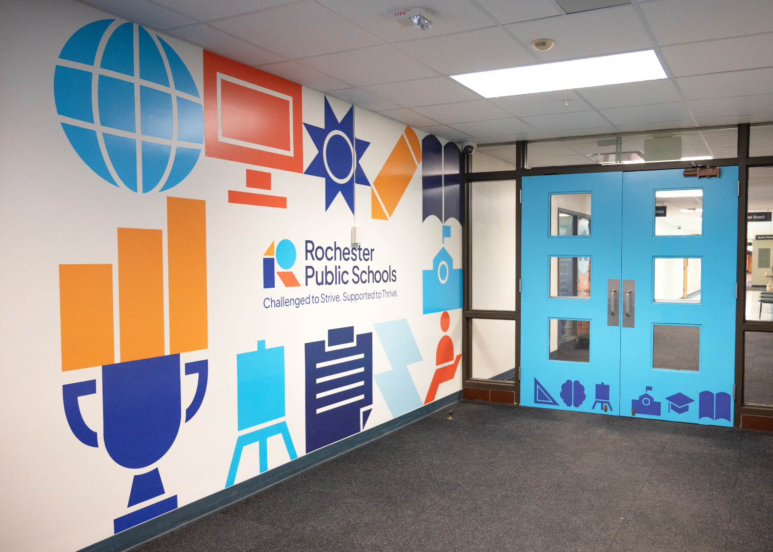 A school hallway featuring a wall mural with colorful icons of school supplies and the Rochester Public Schools logo, leading to a set of bright blue double doors.