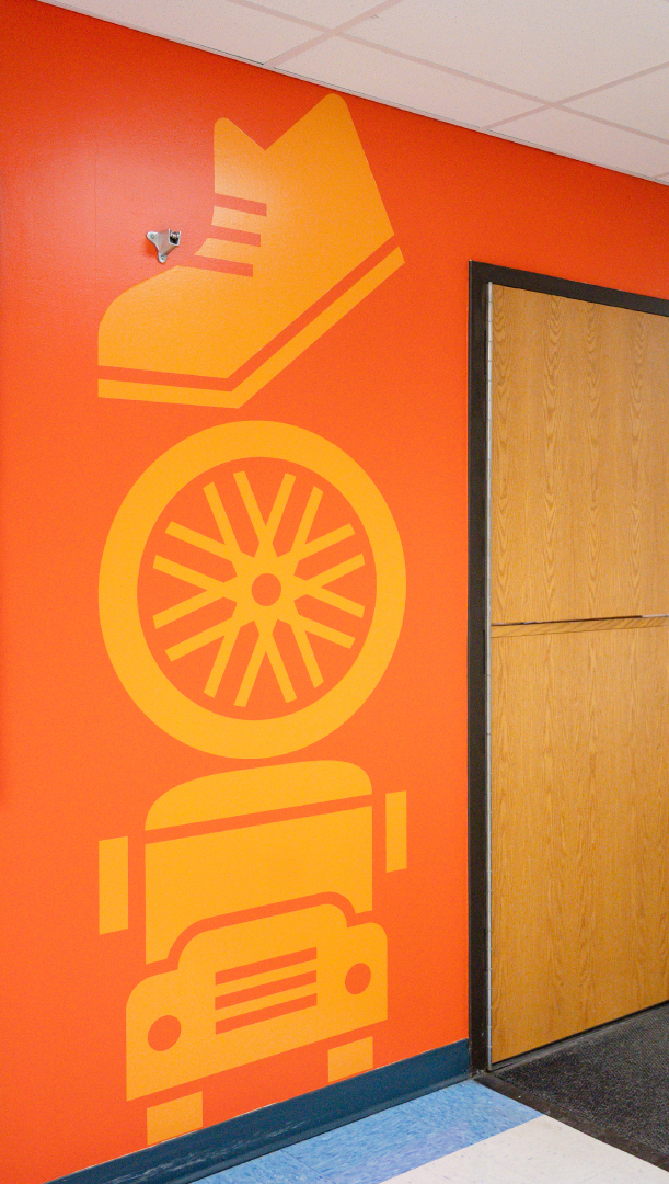 An orange wall decorated with vertical yellow icons of a shoe, a bicycle wheel, and a school bus next to a wooden door.
